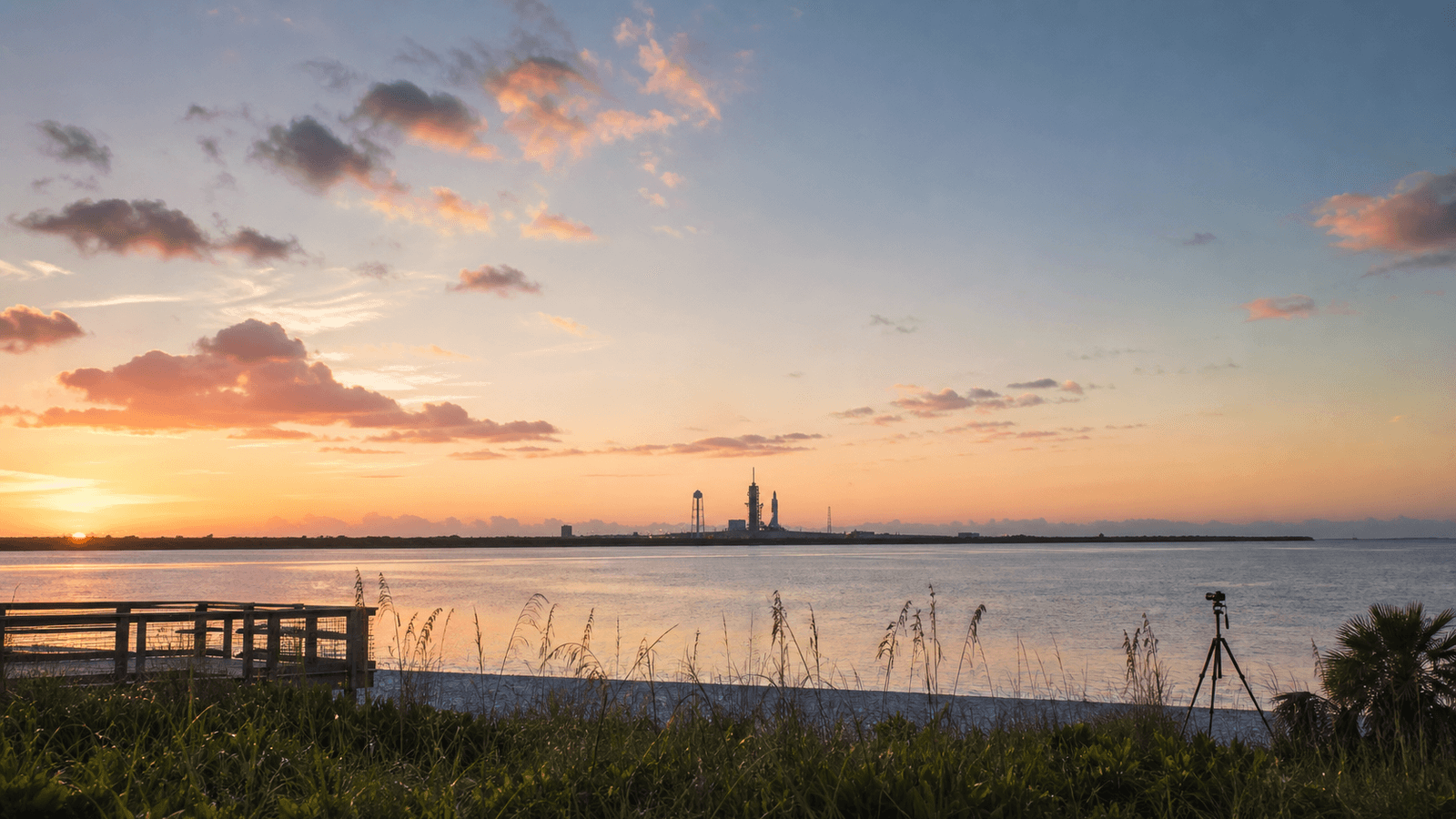Sunset waterfront with a distant launch structure and tripod