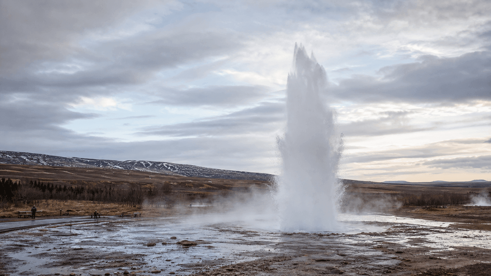 A tall white jet of water rises from a steaming pool, with roped paths and low hills behind it.