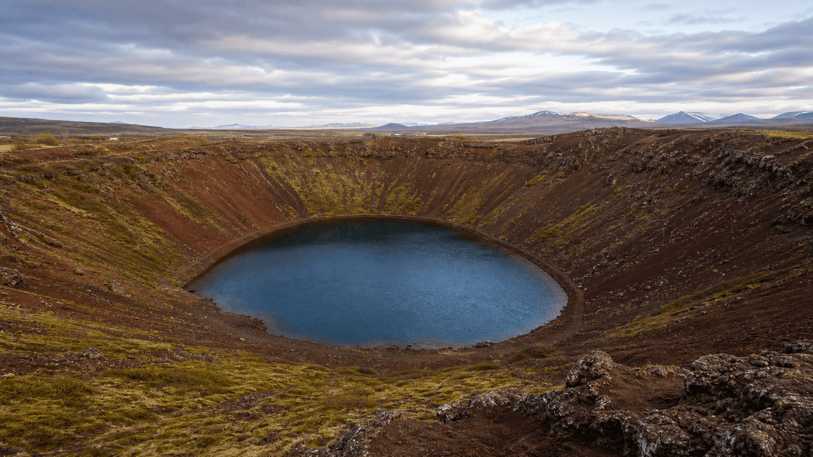 A blue lake fills a reddish-brown crater, with patches of grass and a path around the rim.