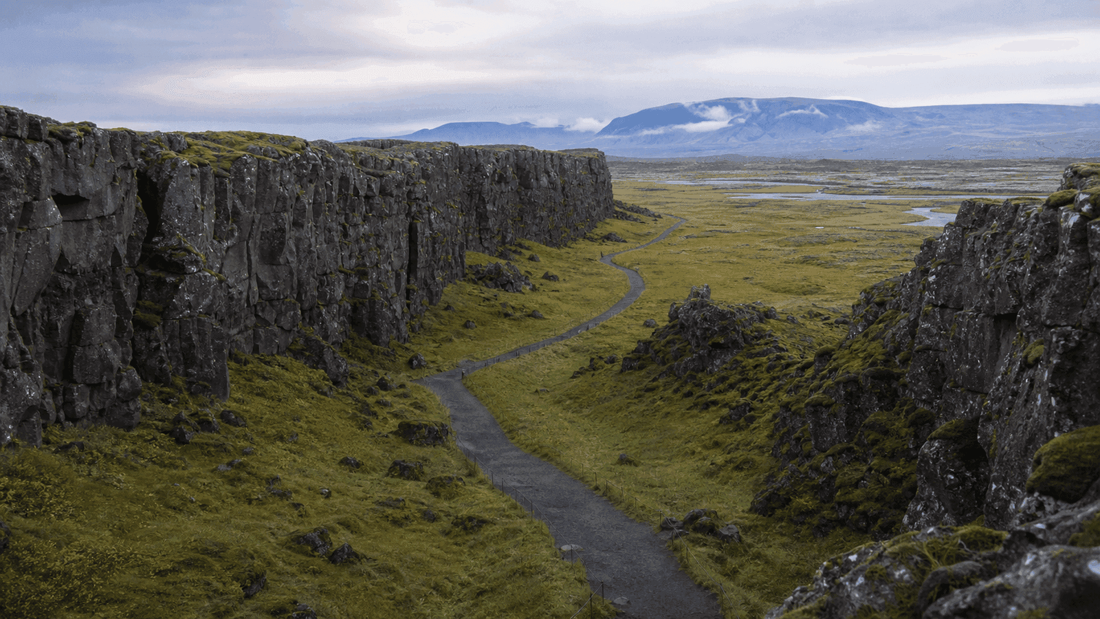 A narrow path between dark rock walls, with grass in the fissure and an open plain beyond.