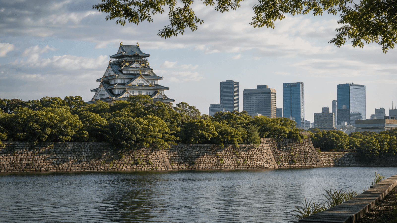 Japanese castle tower behind trees, a stone wall and a moat, with modern high-rises nearby
